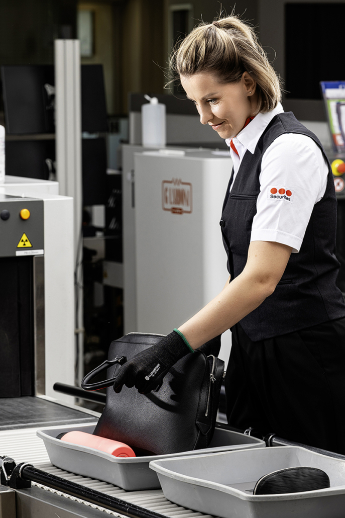 Corporate-Werbefotografie einer Sicherheitsmitarbeiterin bei der Gepäckkontrolle am Flughafen im modernen Employer-Branding- und Kampagnenstil.