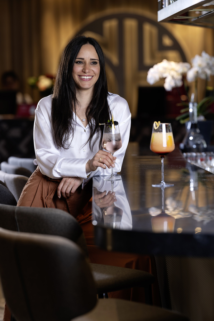 Luxury Hospitality Werbefotografie einer elegant gekleideten Frau an einer stilvollen Hotelbar mit Cocktail im modernen Premium-Lifestyle- und Kampagnenstil