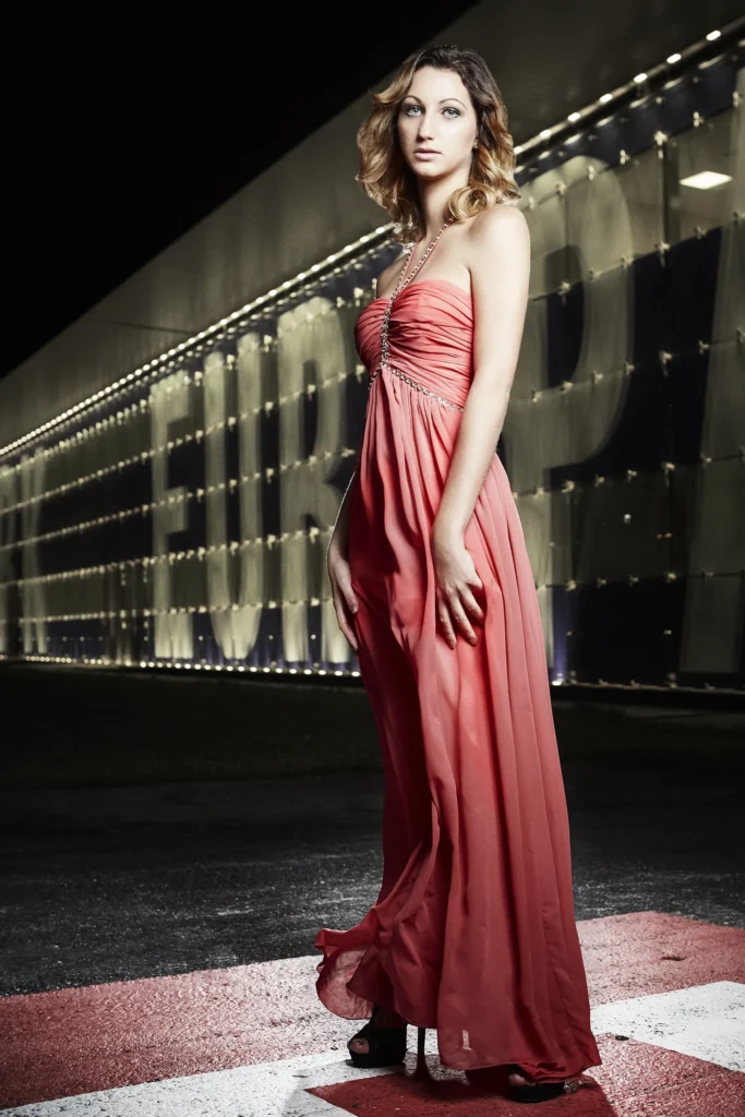 Frau in korallfarbenem Chiffon-Abendkleid mit Neckholder-Ausschnitt und Kristallverzierung steht nachts auf einer beleuchteten Außenanlage mit großformatigen Leuchtbuchstaben im Hintergrund – Magazin-Modefotografie, Fotograf Michael Preschl