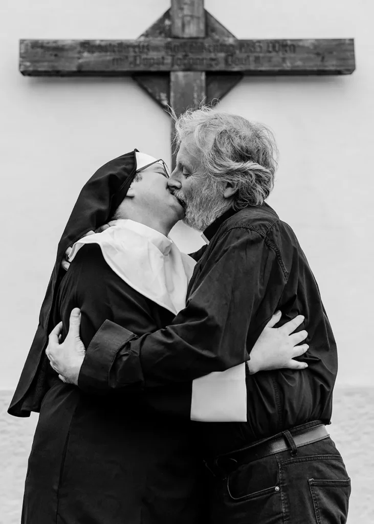 Schwarz-Weiß-Fotografie einer Nonne und eines Priesters in inniger Umarmung vor einem Kreuz. Das provokante und emotional aufgeladene Bild thematisiert Gegensätze, gesellschaftliche Normen und Perspektivenwechsel. Mit dieser Arbeit gewann Claudia Bachinger den Bundeslehrlingswettbewerb der Berufsfotografen Österreich.