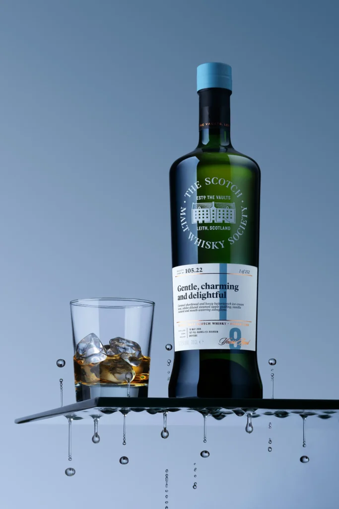 Flasche Scotch Malt Whisky Society Cask 105.22 „Gentle, charming and delightful" auf schwarzer Glasfläche mit Tumbler-Glas auf Eis, Wassertropfen fallen von der Tischkante – Produktfotografie NH Collection, Fotograf Michael Preschl