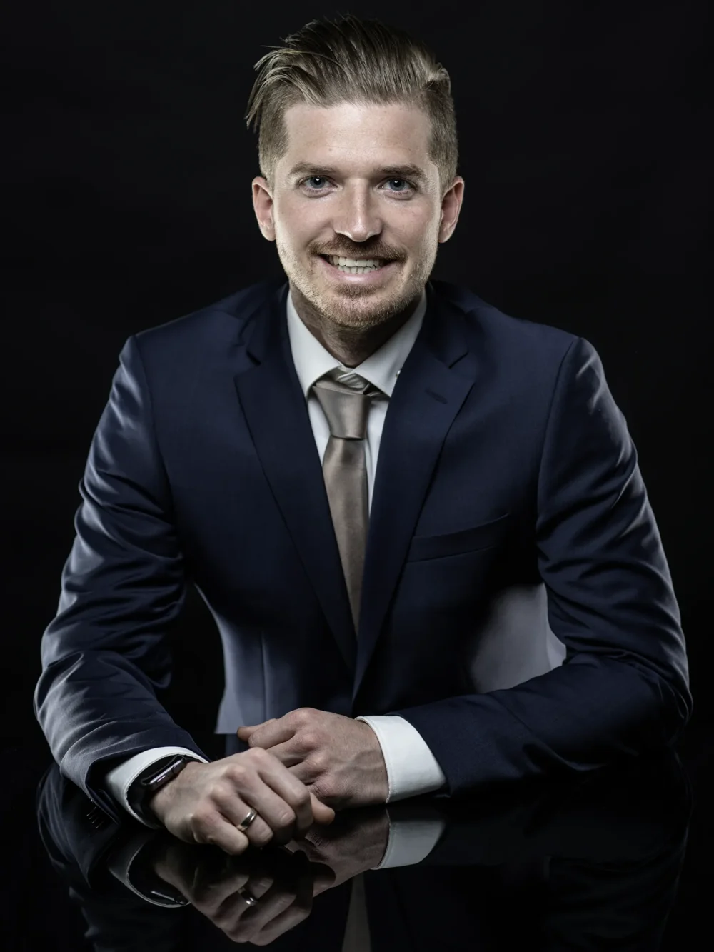 Forbes Austria – Corporate Portrait by Michael Preschl Lächelnder junger Mann in dunkelblauem Anzug mit champagnerfarbener Krawatte lehnt mit verschränkten Händen auf einer Spiegelfläche vor schwarzem Hintergrund – Corporate Portrait Forbes Magazin, Fotograf Michael Preschl