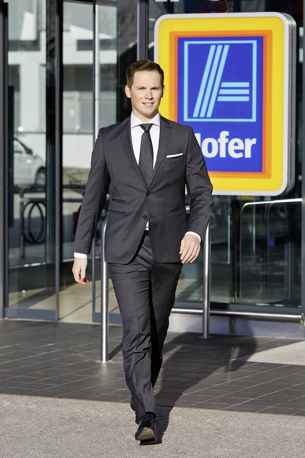 Hofer Austria – Corporate Photography by Michael Preschl Lächelnder Mann in anthrazitfarbenem Anzug mit schwarzer Krawatte und weißem Einstecktuch läuft auf die Kamera zu, im Hintergrund der Hofer-Markeneingang mit gelbem Logo – Corporate Brand Visual, Fotograf Michael Preschl