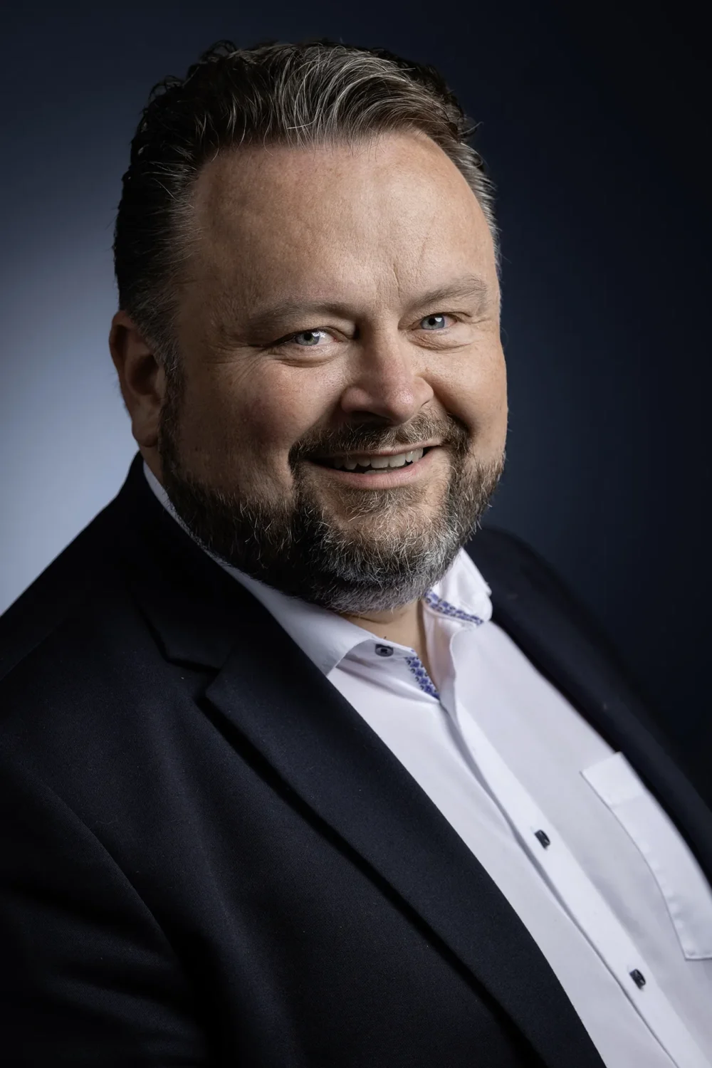 Mario Friedl — Portrait for Forbes Austria by Michael Preschl Mario Friedl lächelt direkt in die Kamera, trägt schwarzen Anzug und weißes Hemd mit blauer Stickerei, vor dunkelblauem Studiohintergrund – Magazin-Portrait Forbes Austria, Fotograf Michael Preschl