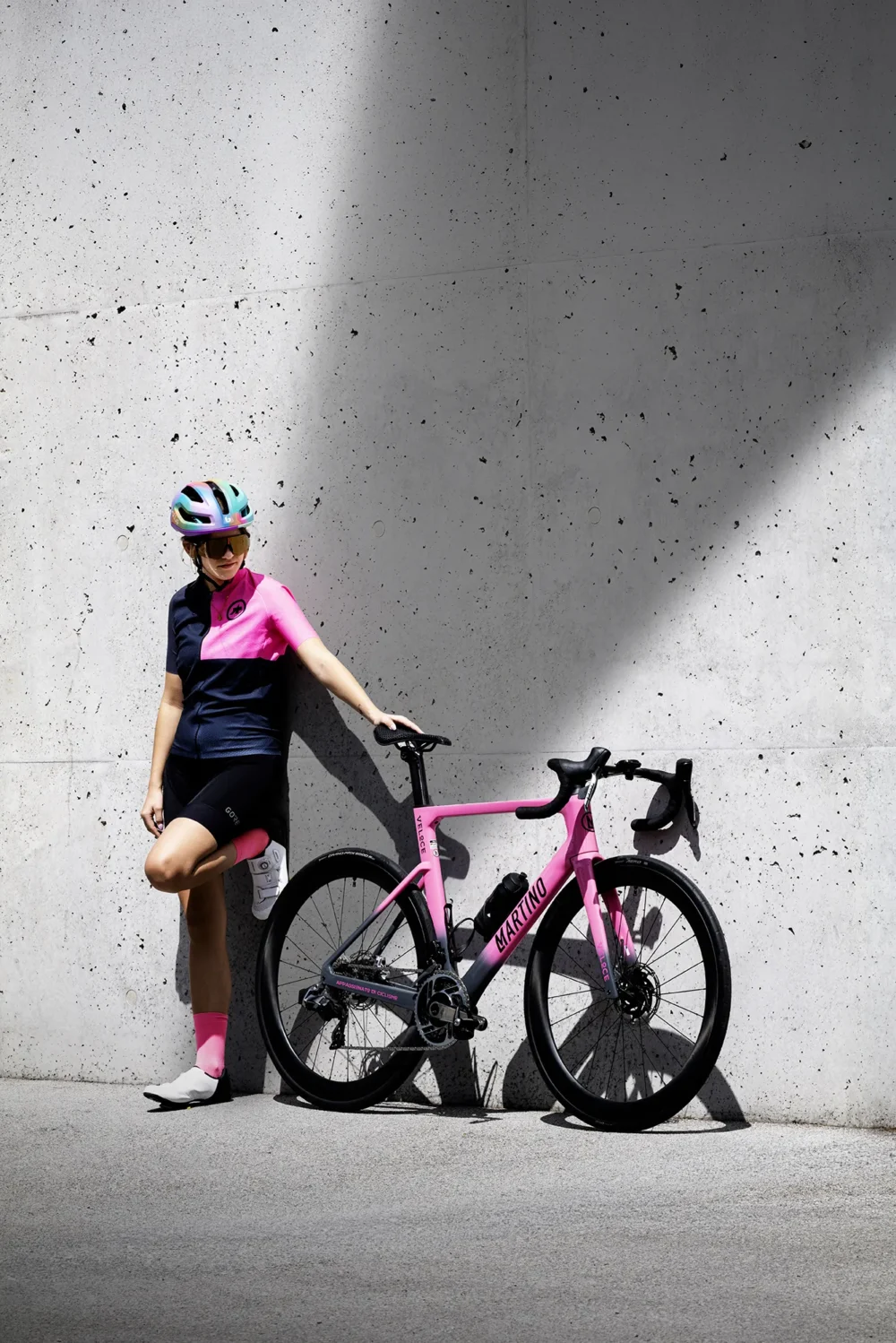 Built to race. Worn to win. Martino Cycling — Shot by Michael Preschl Radfahrerin in navy-pinkfarbenem Assos-Trikot, schwarzen Bib-Shorts, pinkfarbenen Socken und irisierendem Helm lehnt neben einem pinkfarbenem Martino-Rennrad an einer Betonwand mit hartem Schattenwurf – Werbekampagne Martino Cycling, Fotograf Michael Preschl