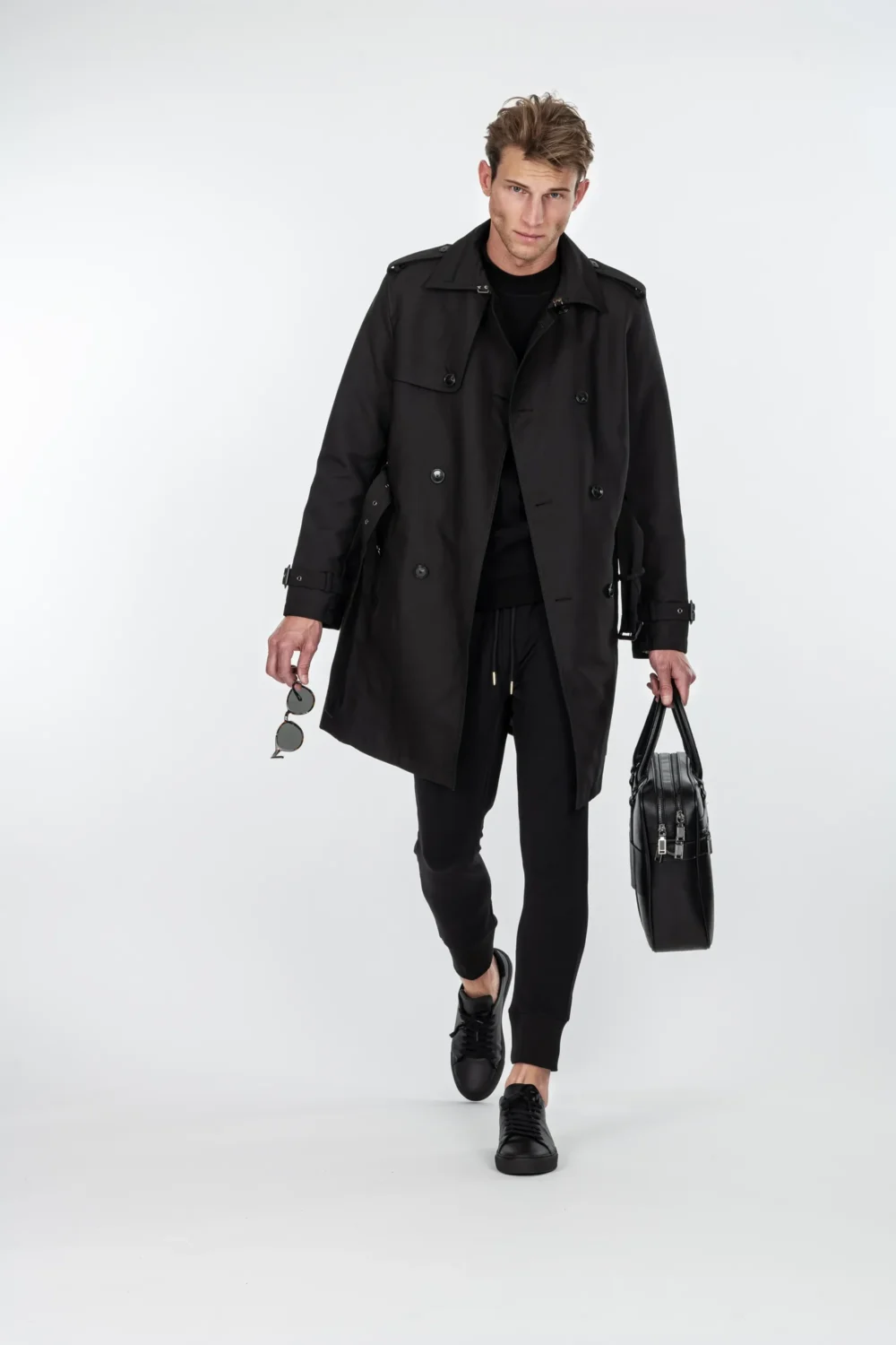 Männliches Model in schwarzem Doppelreiher-Trenchcoat, schwarzem Sweater, Jogger-Hose und schwarzen Sneakers läuft auf weißem Studiohintergrund, Sonnenbrille in der rechten Hand, schwarze Leder-Laptoptasche in der linken – Modefotografie ICAN, Fotograf Michael Preschl