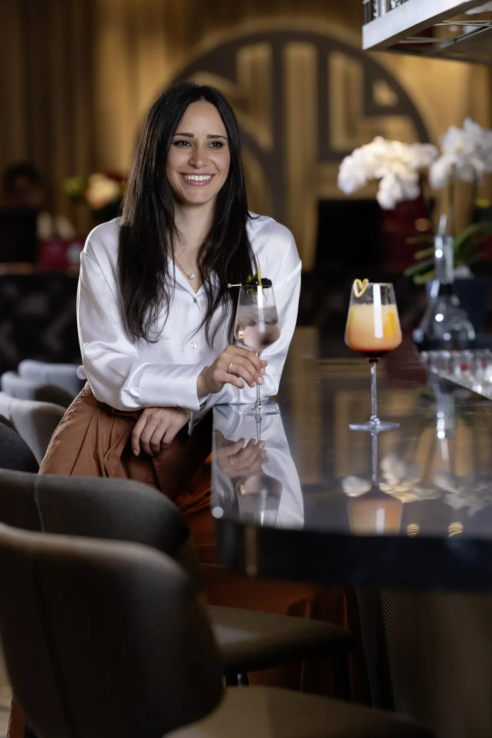 Lächelnde Frau mit langen dunklen Haaren in weißer Seidenbluse und cognacfarbener Hose lehnt an einer Hotelbar, hält ein Cocktailglas, im Hintergrund das NH Hotels Logo – Corporate Brand Visual, Fotograf Michael Preschl