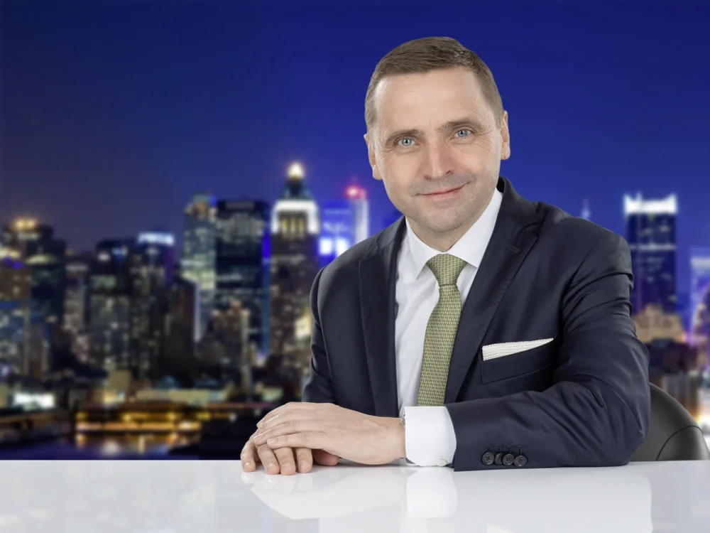 RT Reisen – Corporate Portrait by Michael Preschl Lächelnder Mann in dunkelblauem Anzug mit olivgrüner Krawatte und weißem Einstecktuch sitzt an einem weißen Tisch vor einer unscharfen nächtlichen Stadtsilhouette – Corporate Portrait RT Reisen, Fotograf Michael Preschl