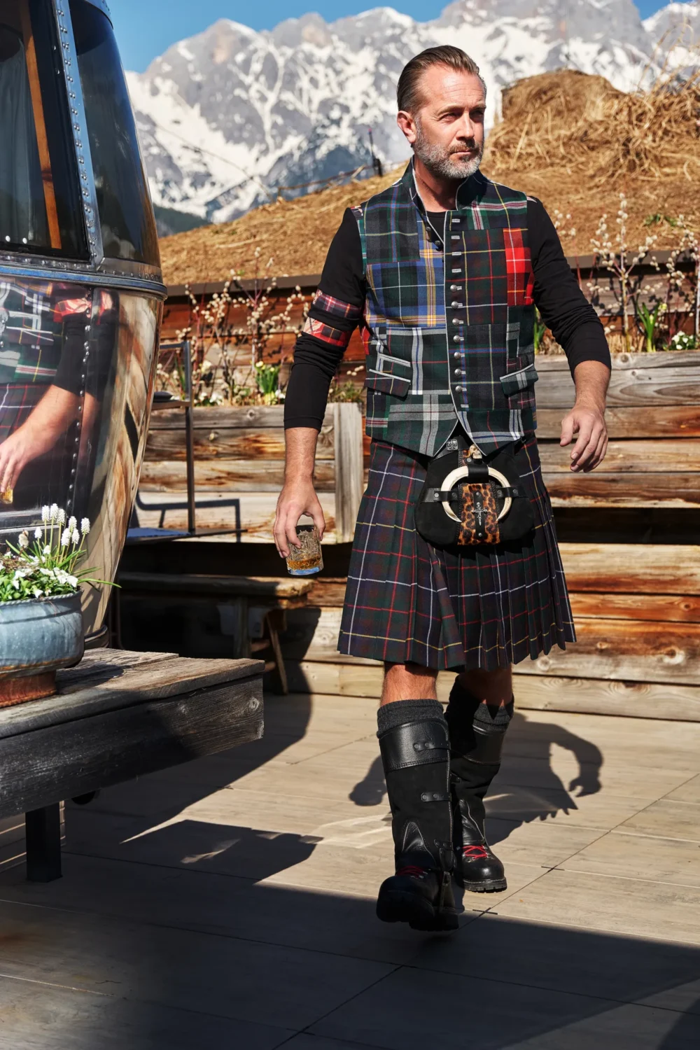 Andreas Mittermaier in Rettl 1886 Tartan-Kilt mit passendem Patchwork-Gilet, schwarzem Langarmshirt und Lederstiefeln auf einer Almterrasse vor verschneitem Bergpanorama, Whisky-Glas in der Hand