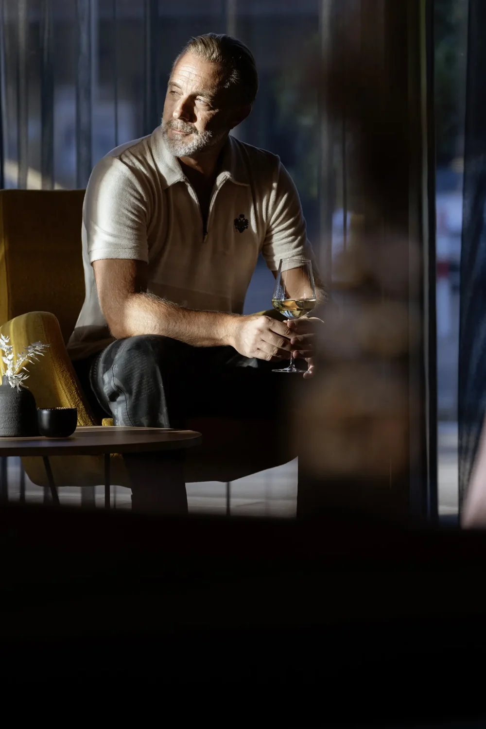 Stille Stärke. Andreas Mittermaier – Portrait by Michael Preschl Andreas Mittermaier sitzt in einem senfgelben Sessel in einer gedimmten Lounge-Atmosphäre, hält ein Weißweinglas, trägt ein cremefarbenes Polo-Shirt mit Wappenadler-Emblem – Magazin-Portrait, Fotograf Michael Preschl