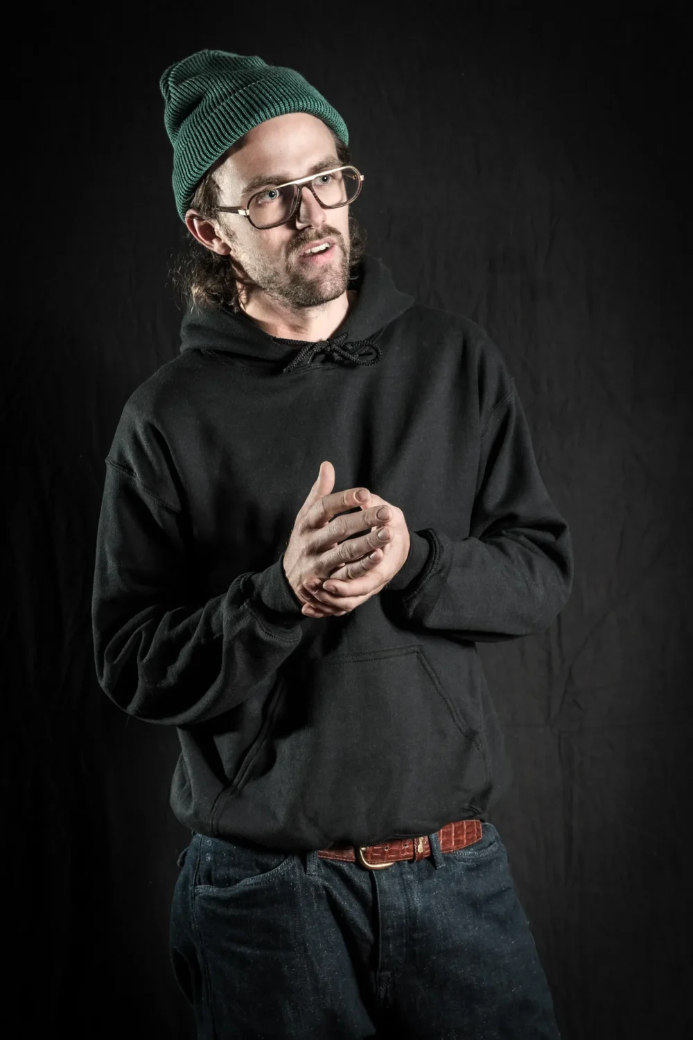 Felix Schelhorn mit grüner Strickmütze, eckiger Hornbrille und schwarzem Hoodie vor dunklem Studiohintergrund – Corporate Portrait für Rolling Pin Magazin, Fotograf Michael Preschl