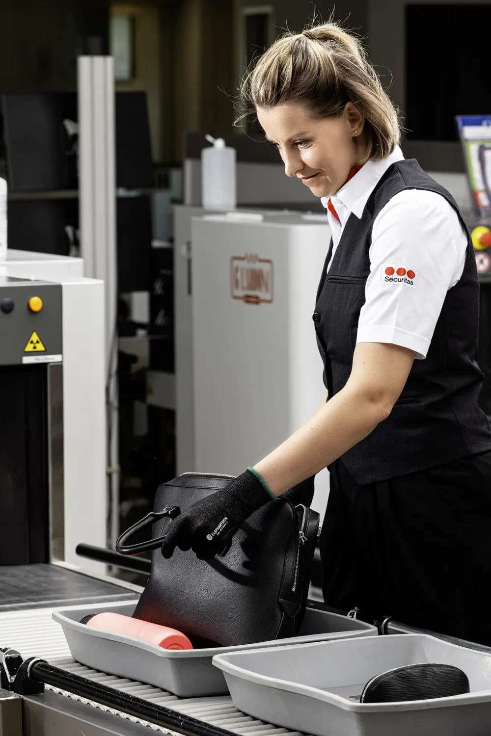Sicherheit mit Haltung. Securitas — Corporate Photography by Michael Preschl Lächelnde Securitas-Mitarbeiterin in weißem Hemd, schwarzer Weste und schwarzen Elephant Skin Schutzhandschuhen kontrolliert eine schwarze Tasche am Gepäckscanner einer Sicherheitskontrolle – Corporate Kampagnenfotografie, Fotograf Michael Preschl