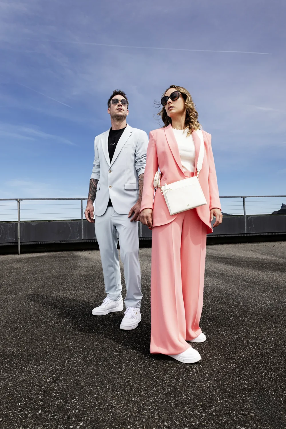 Power Couple. Strellson SS-Collection — Shot by Michael Preschl Mann im hellblauen Strellson-Anzug mit schwarzem T-Shirt und Aviator-Sonnenbrille neben Frau im rosafarbenen Blazer-Set mit weißer Umhängetasche, beide auf einem Rooftop vor blauem Himmel