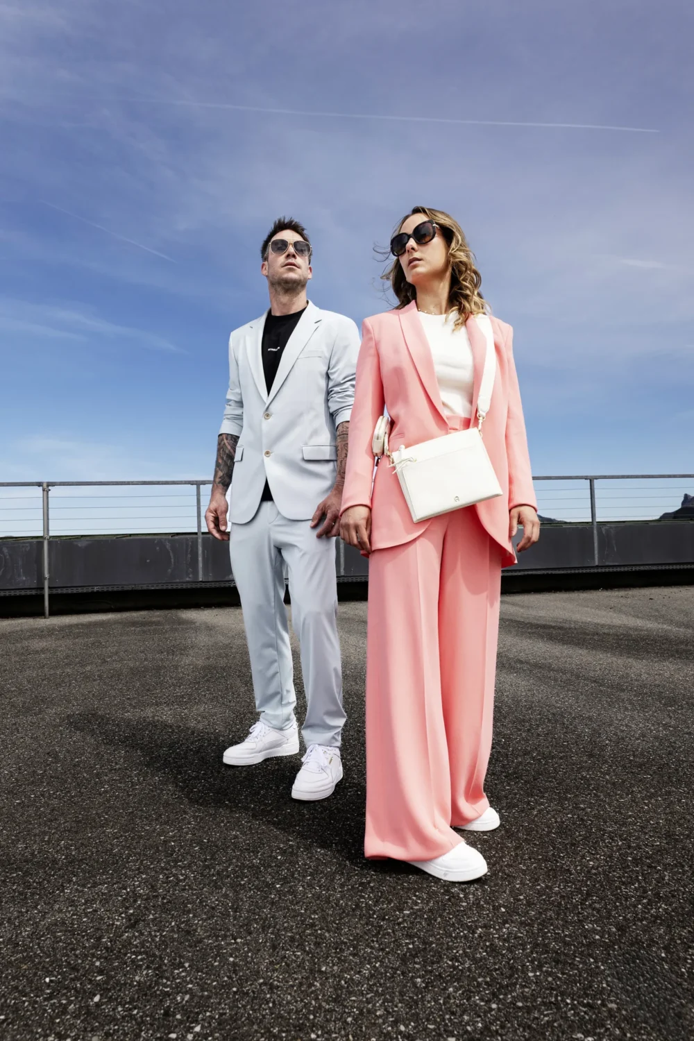 Mann im hellblauen Strellson-Anzug mit schwarzem T-Shirt und Aviator-Sonnenbrille neben Frau im rosafarbenen Blazer-Set mit weißer Umhängetasche, beide auf einem Rooftop vor blauem Himmel