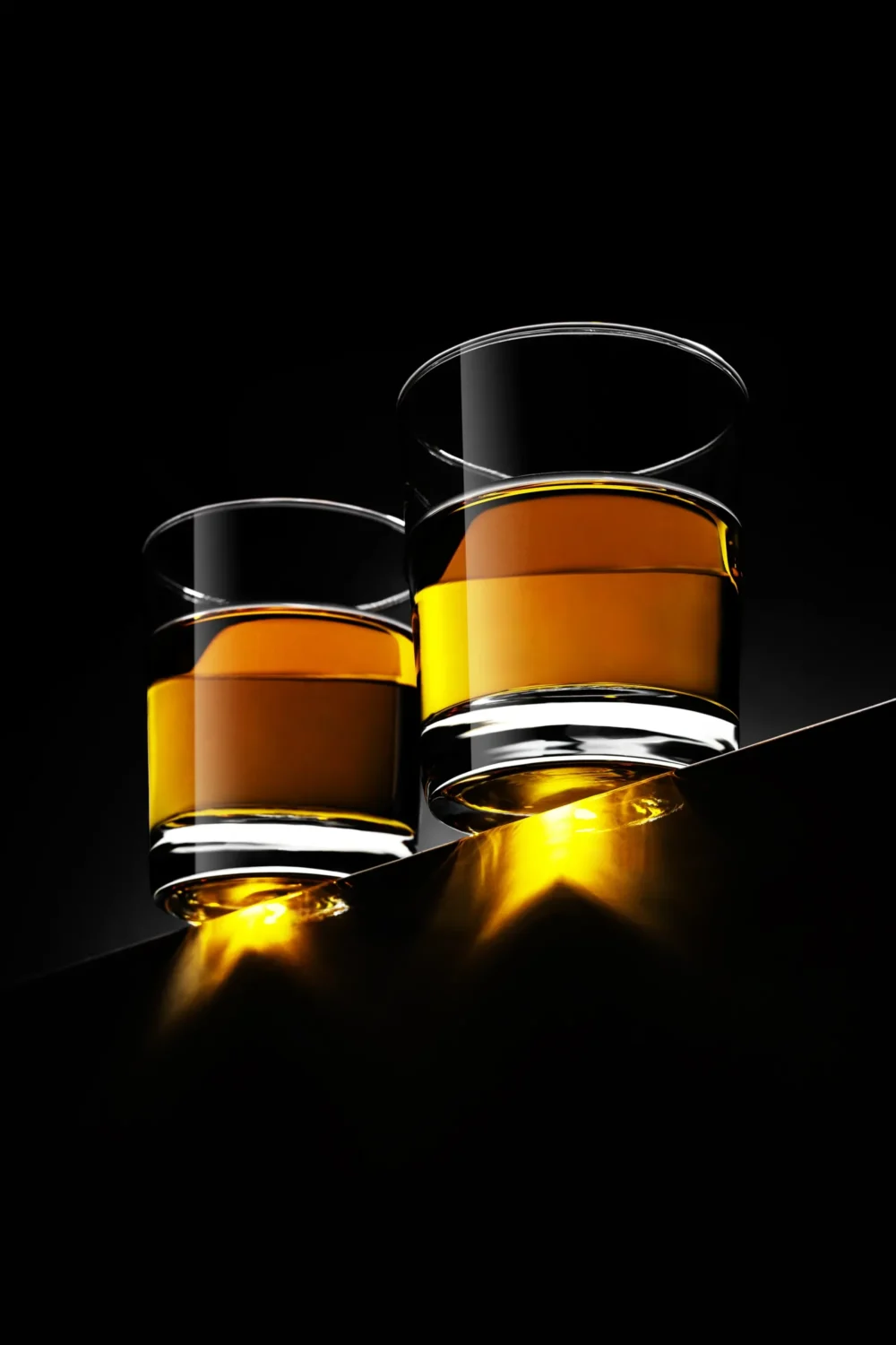 Pure Gold. Whisky Product Photography by Michael Preschl Zwei Tumbler-Gläser mit Whisky auf schwarzer Spiegelfläche vor dunklem Hintergrund, goldgelbe Lichtreflexe durch den Glasboden – Produktfotografie Michael Preschl